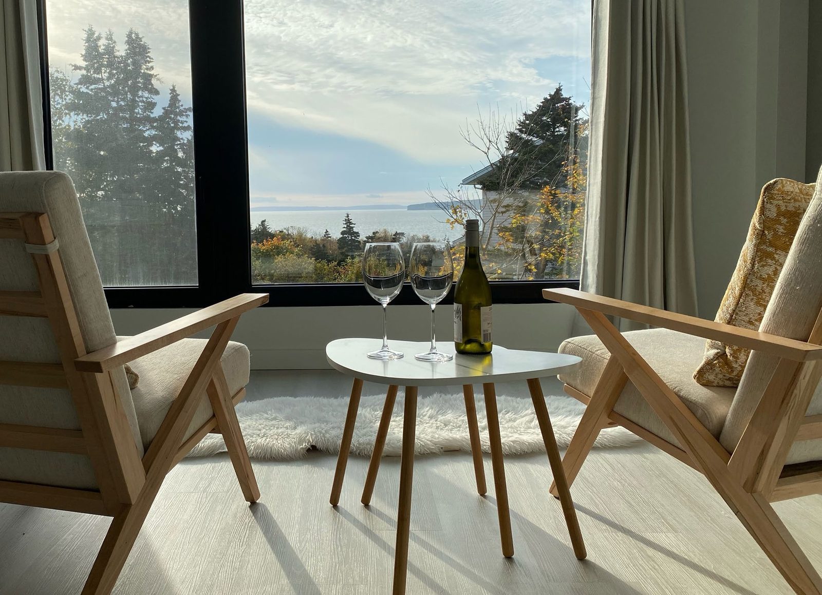 A pair of light wood armchairs framing a small white table set with two wine glasses, in front of a large window overlooking the Newfoundland coastline.