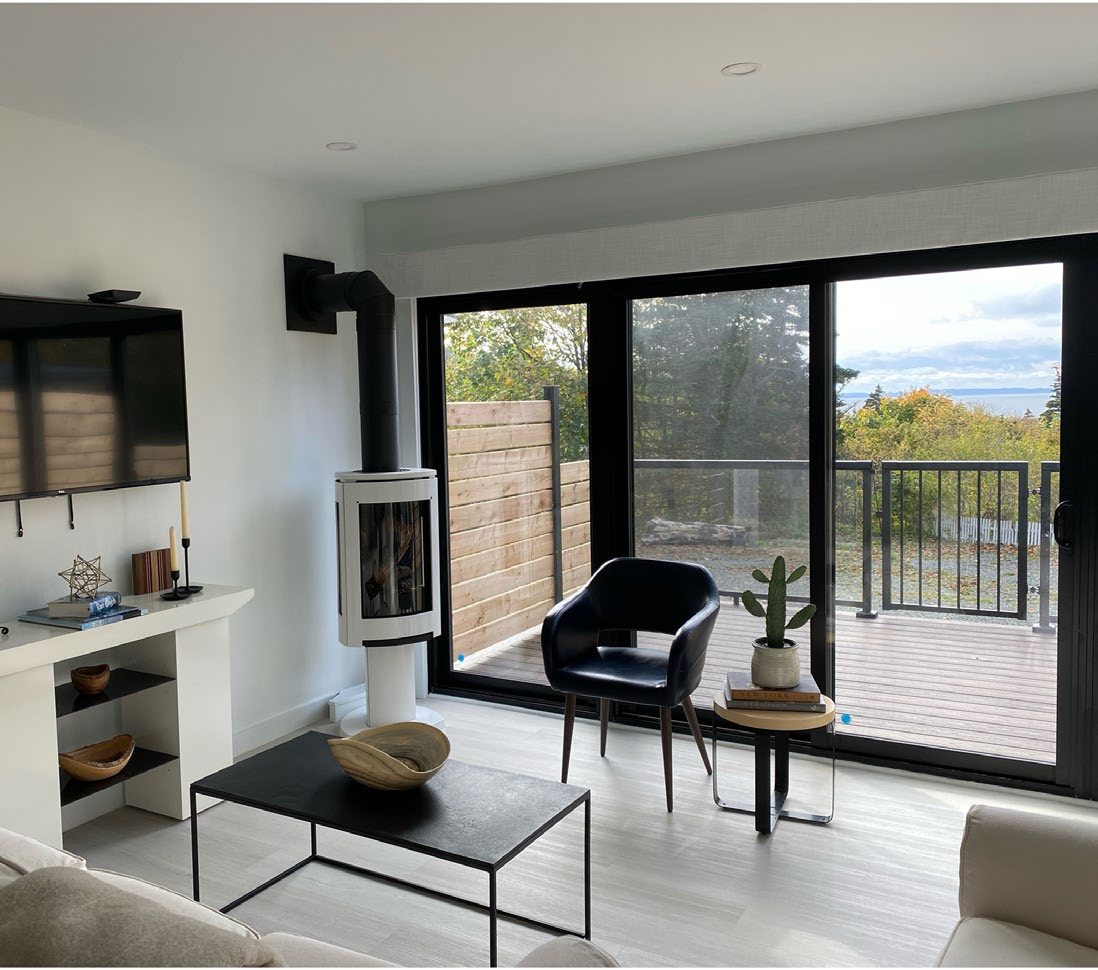A bright, modern living room with a small wood stove, a black-framed dining chair, and floor-to-ceiling glass doors opening onto a deck with an ocean view.