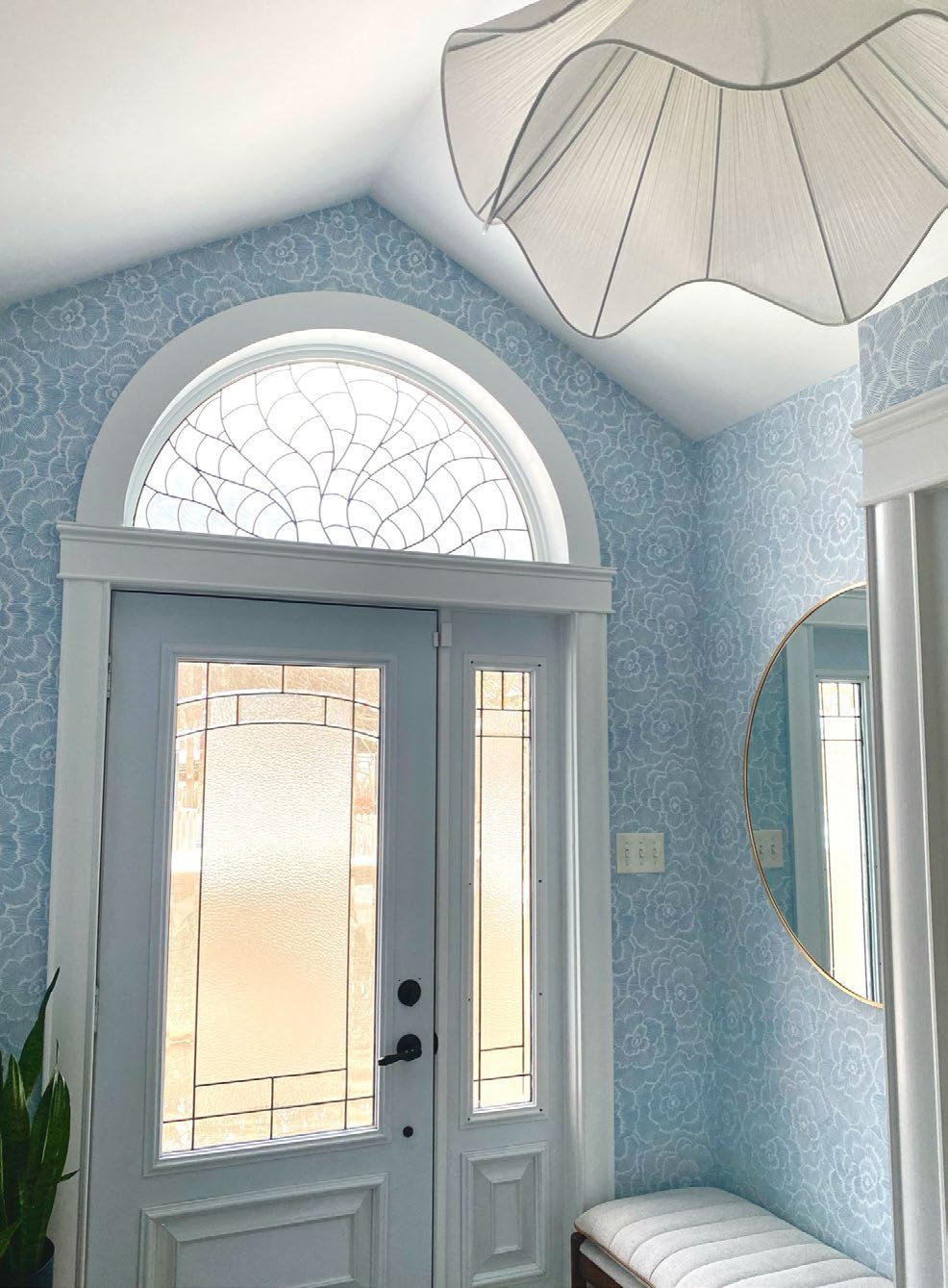 An entryway with soft blue floral wallpaper, an arched leaded-glass transom window, and a white scallop-shaped pendant light.