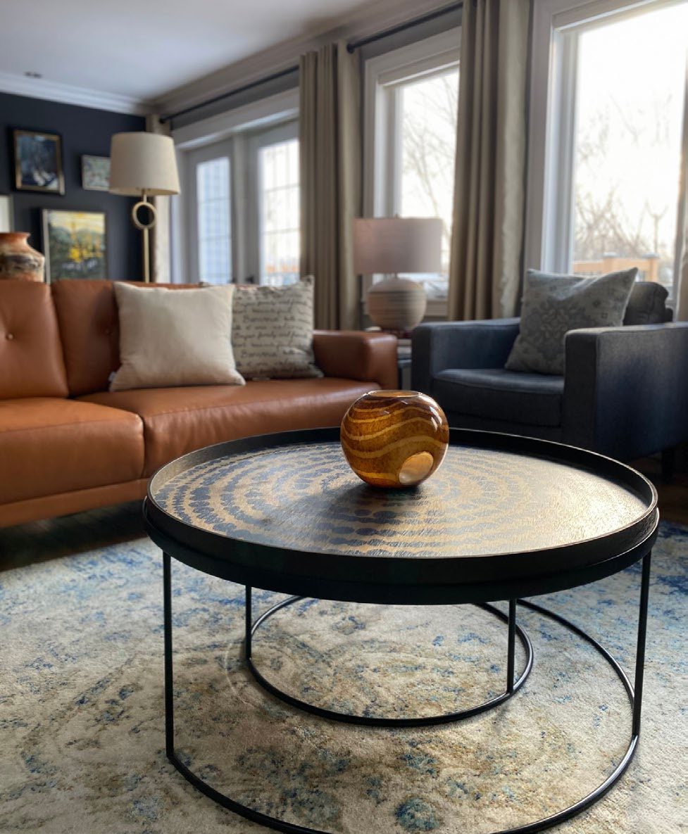 A classic living room with a tan leather sofa, navy accent chair, round metal-and-stone coffee table, and patterned area rug.