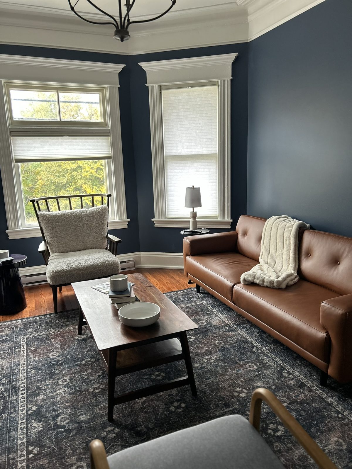 A traditional living room with deep navy walls, a tan leather sofa with a chunky cream throw, a cream bouclé accent chair, dark wood coffee table, and a patterned area rug.