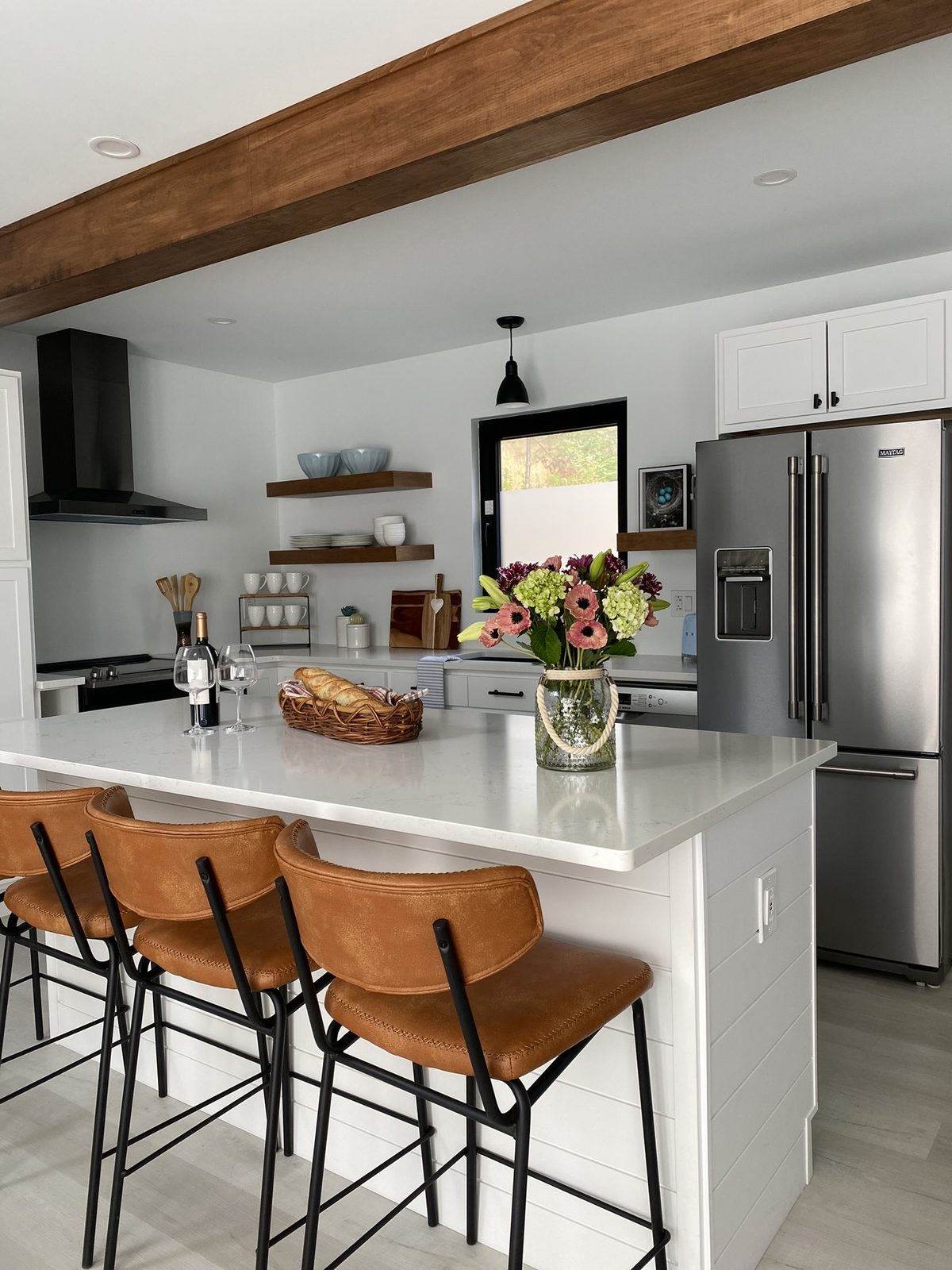 An open-concept kitchen with an exposed wood beam, white shaker cabinets, walnut floating shelves, black range hood, and a large island with tan leather counter stools and fresh flowers.