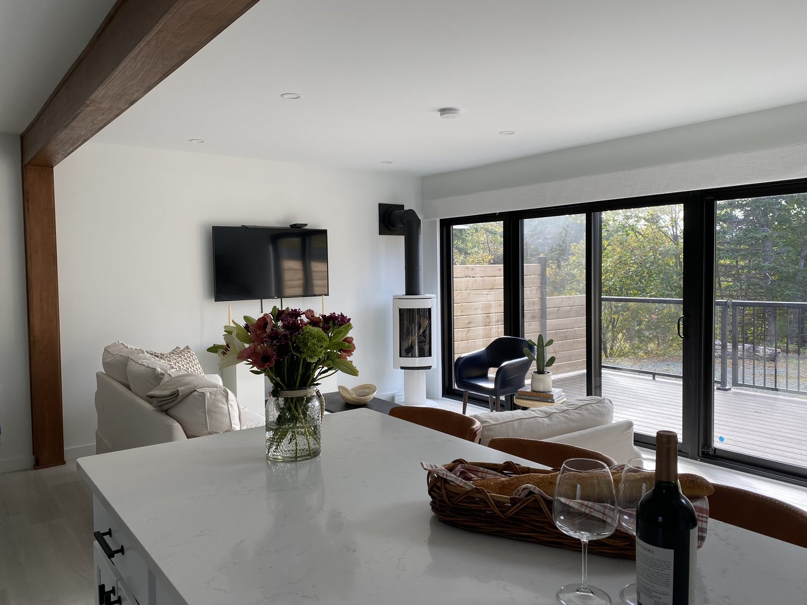 An open-concept living and kitchen area with an exposed wood beam, white sofa, fresh flowers on a quartz island, a freestanding white woodstove, navy accent chair, and sliding doors to a deck with woodland views.