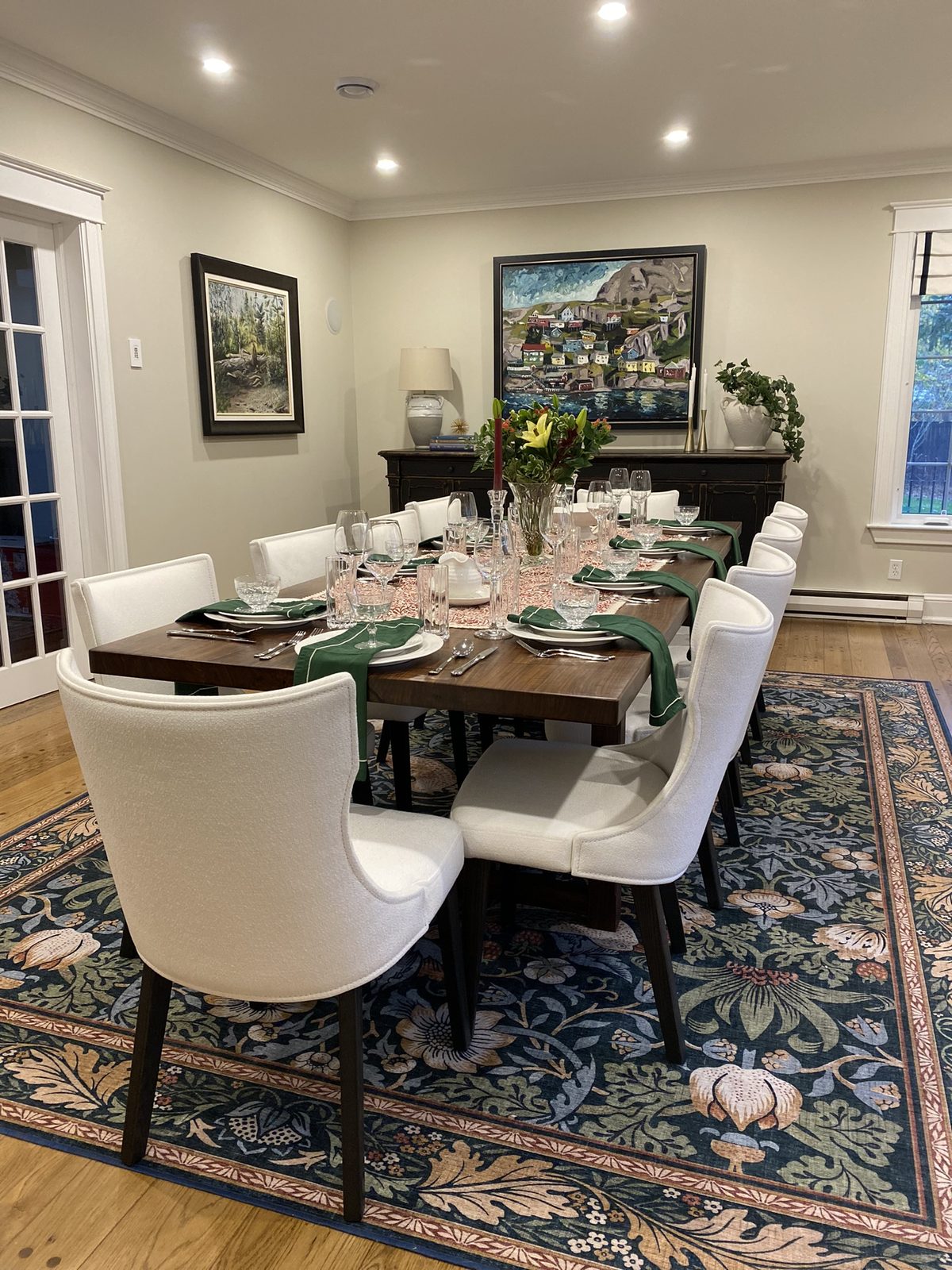 A formal dining room with a wood table set for eight, white parsons chairs paired with bouclé end chairs, green linen napkins, a landscape painting above a sideboard, and a floral-patterned rug.