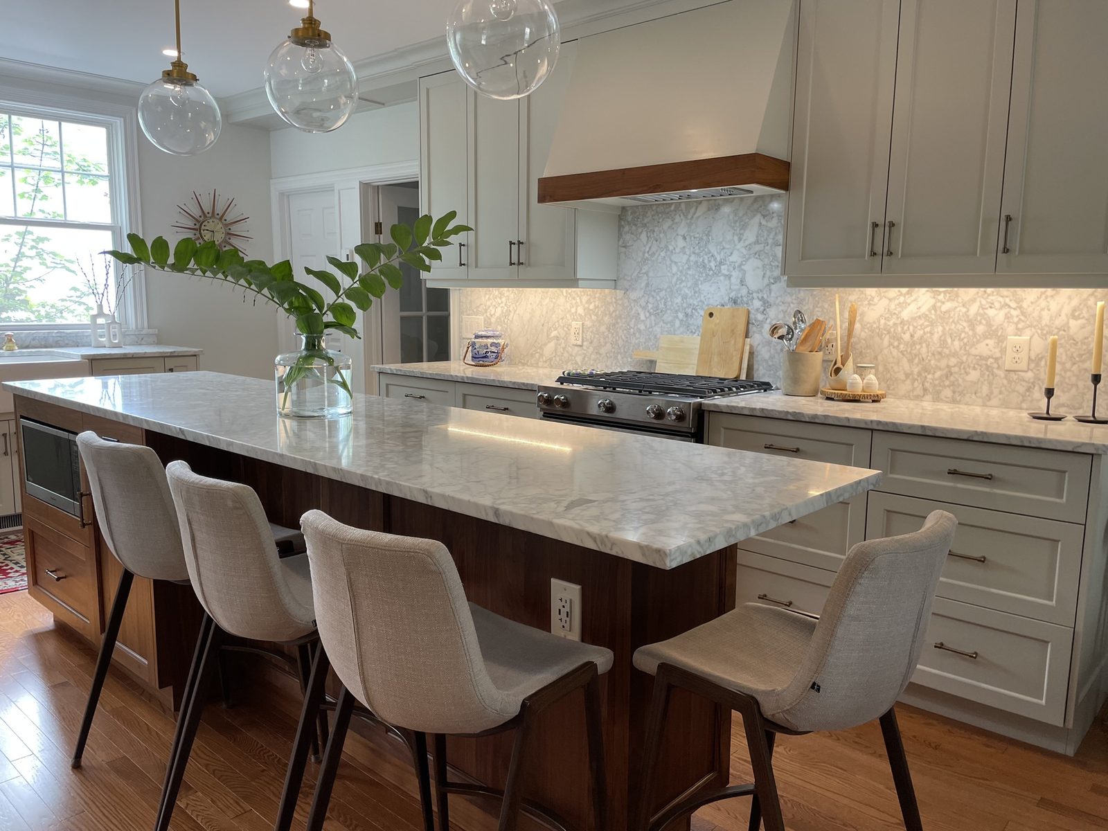 A spacious kitchen with light grey shaker cabinets, full marble backsplash and counters, a walnut-trimmed range hood, three glass globe pendants, and an island with light grey upholstered stools.