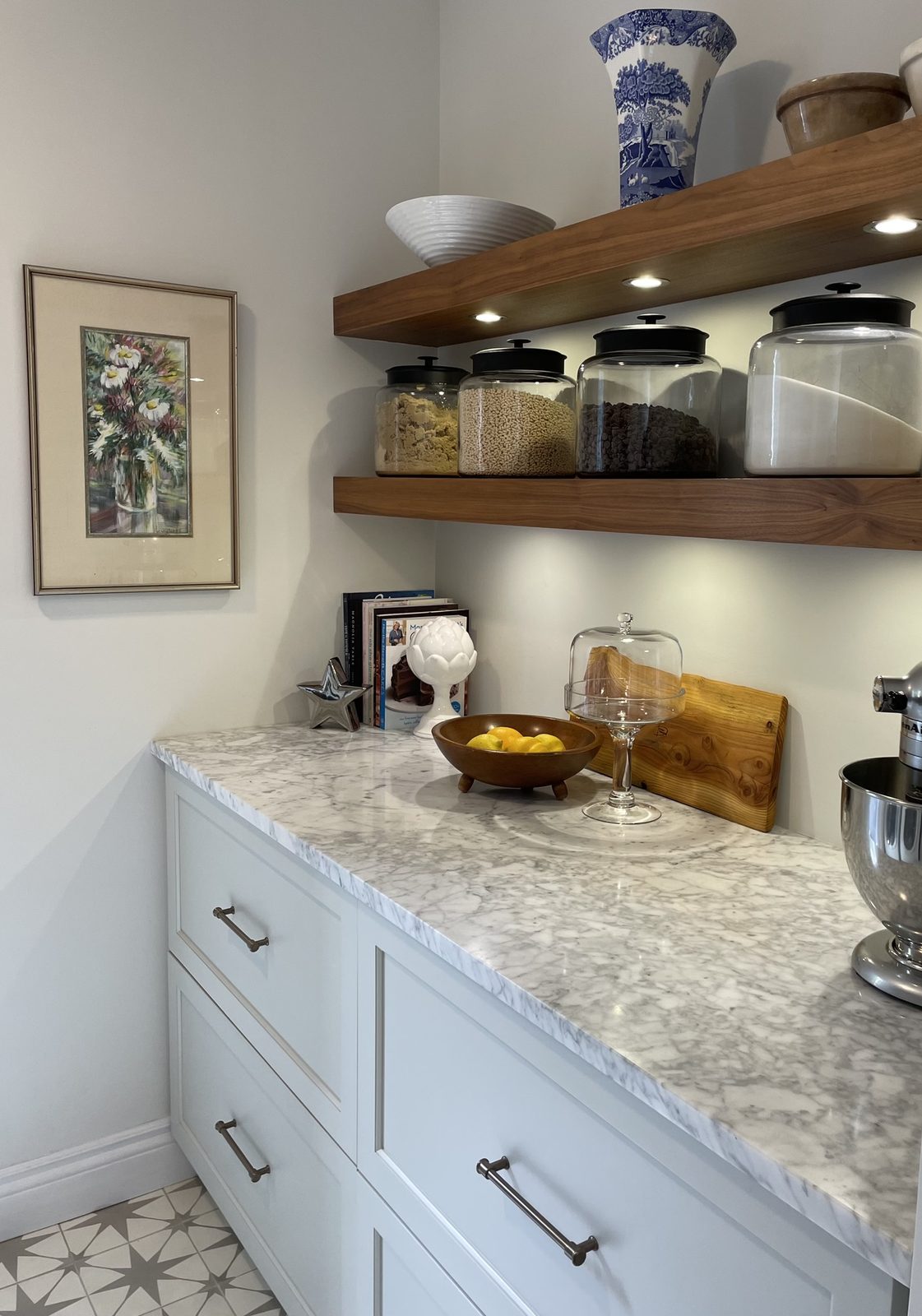 A butler's pantry with walnut floating shelves displaying glass canisters and a blue-and-white ginger jar, marble counters, light blue base cabinets, patterned tile floor, and a framed floral watercolor.