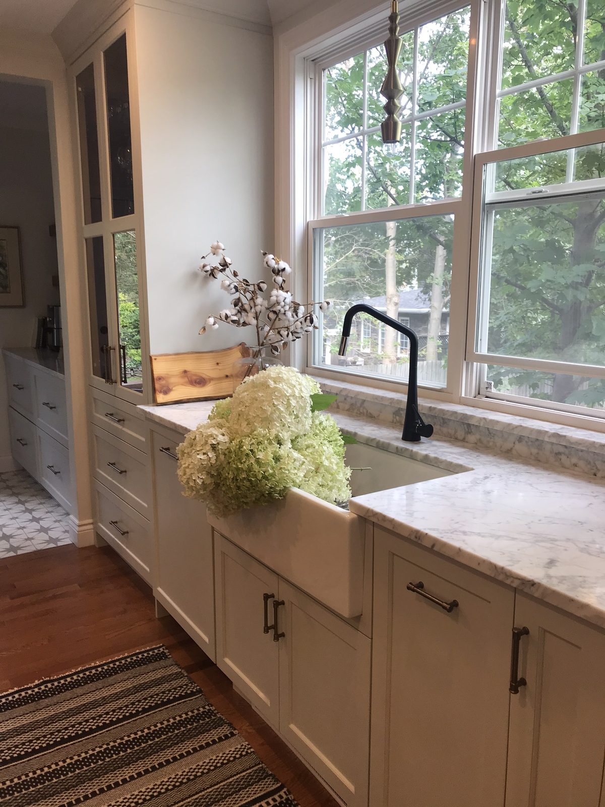 A bright farmhouse kitchen with white shaker cabinets, a deep apron-front sink, marble countertops, and a vase of hydrangeas in front of a large window overlooking trees.