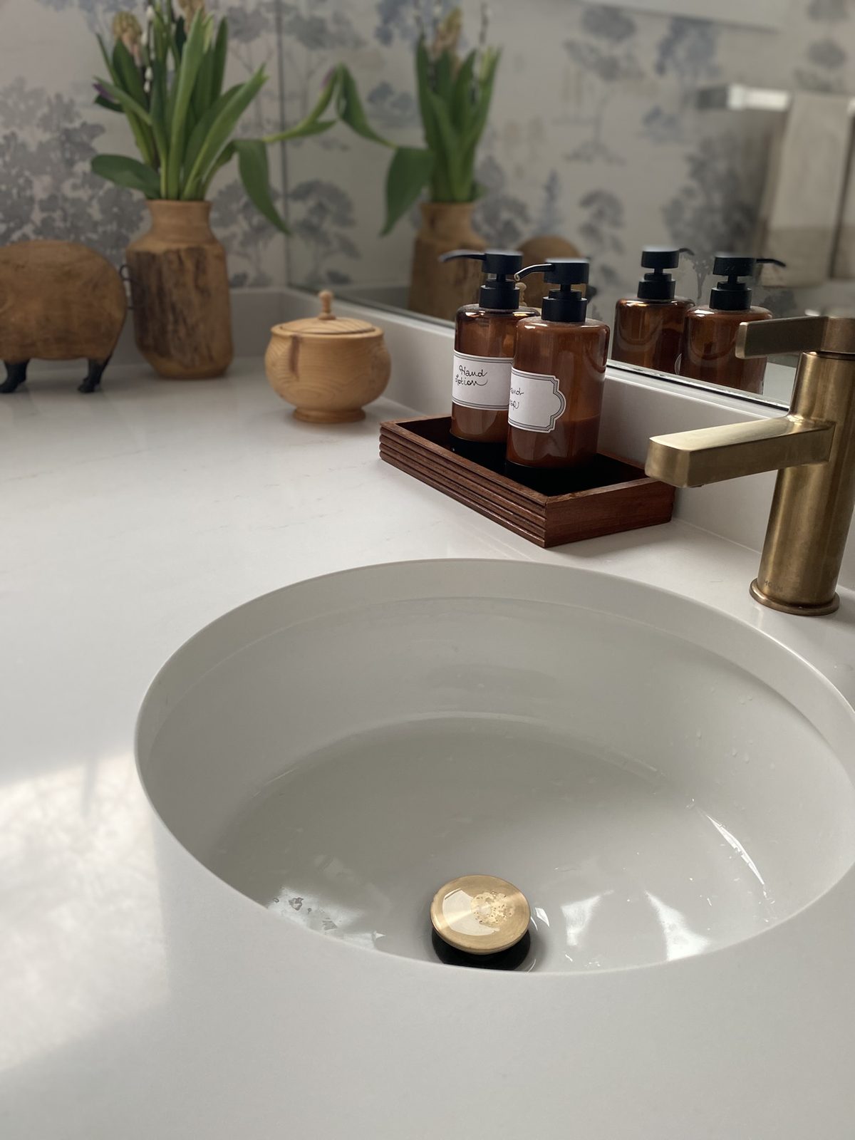 A close-up powder room vignette with a white quartz countertop, brushed brass faucet, oval undermount sink, amber glass soap bottles in a wooden tray, and hyacinths in carved wood vessels against blue toile wallpaper.