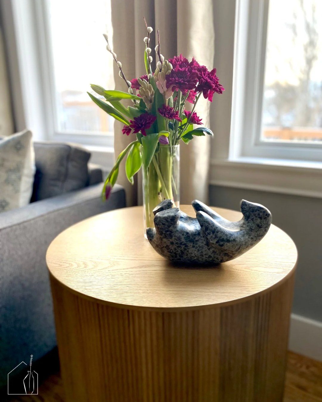 A round fluted oak side table styled with a sculptural stone vessel and a clear glass vase of dried pink flowers and pussy willows, beside a soft gray sofa.