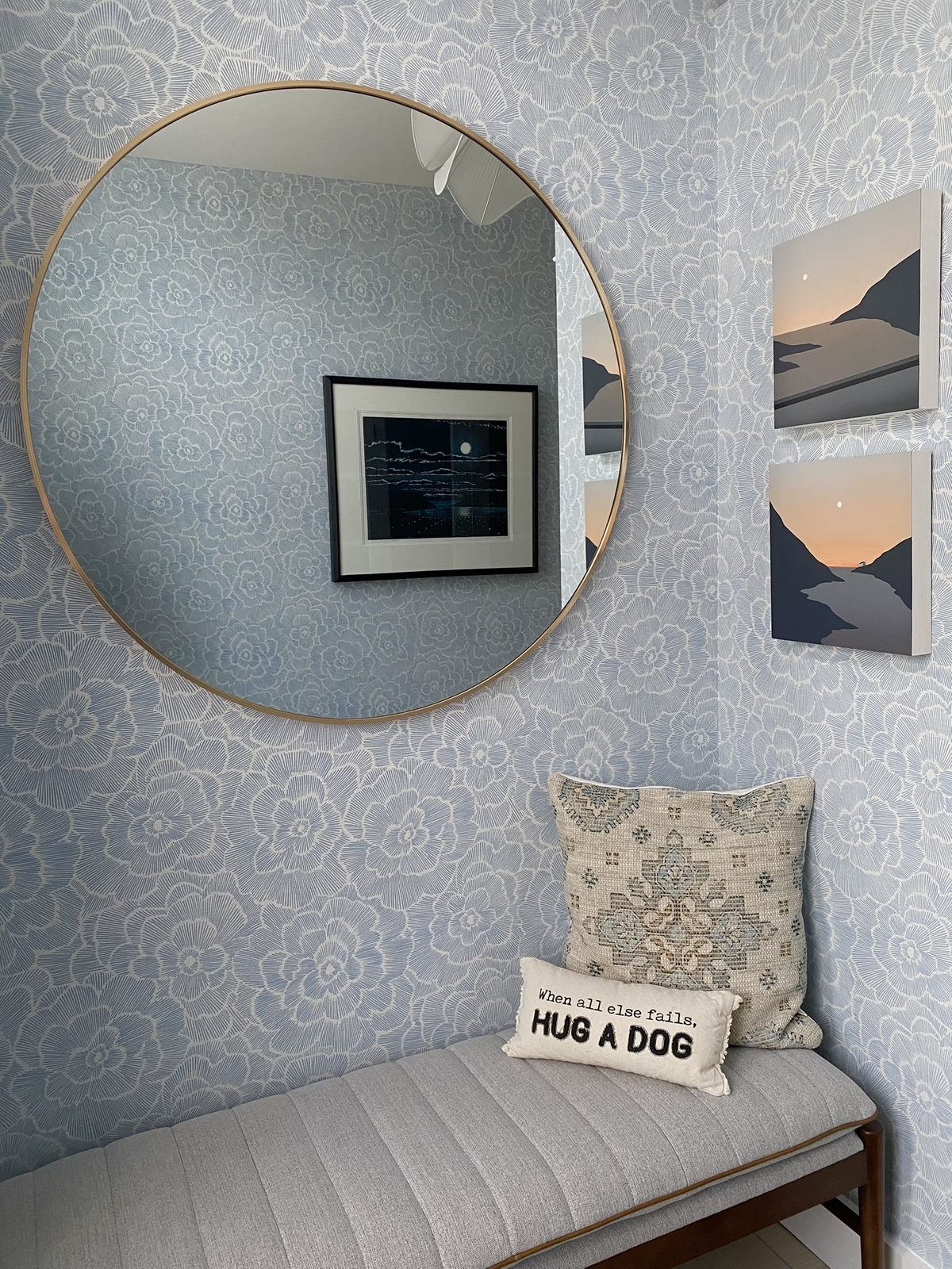 A styled corner featuring a large round brass mirror on blue floral wallpaper, two sunset-toned art prints, and a gray upholstered bench with patterned pillows.